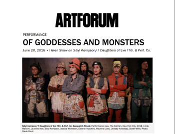 A group of women clad in aprons & beanies in assorted garish colors and patterns, and headlamps, stand in dim theatrical light, at close range, with arms crossed, in the midst of forming words, seemingly in unison.