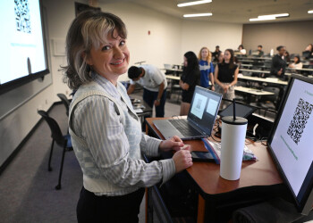 Dr. Zwierzchowski stands at a podium smiling while students can be seen in the background engaging in a s